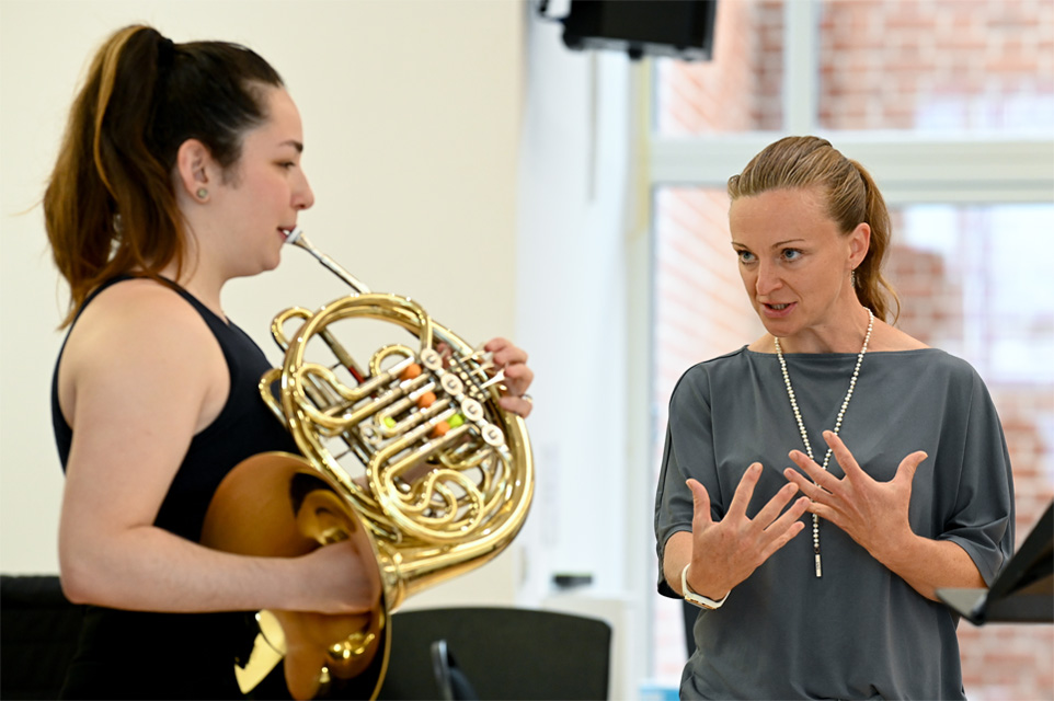A student performing on the French Horn in front of Annamia Larsson, a women with light hair, guiding her performance on stage.